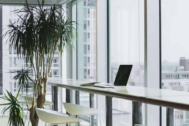 a long desk with laptop looking out a building next to small potted plant